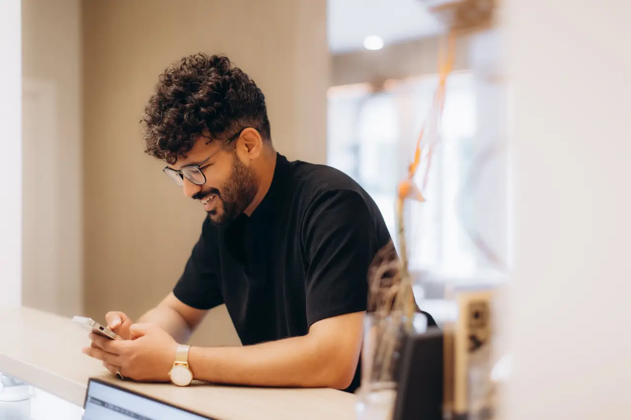 A man smiling at his phone while leaning on a kitchen bench