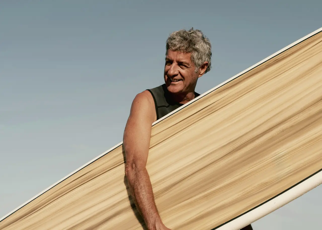 A smiling grey-haired surfer carrying a long wooden surfboard under a blue sky