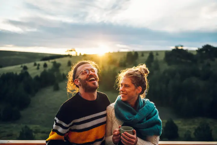 A couple laughing together with a hot drink on a hillside at sunset