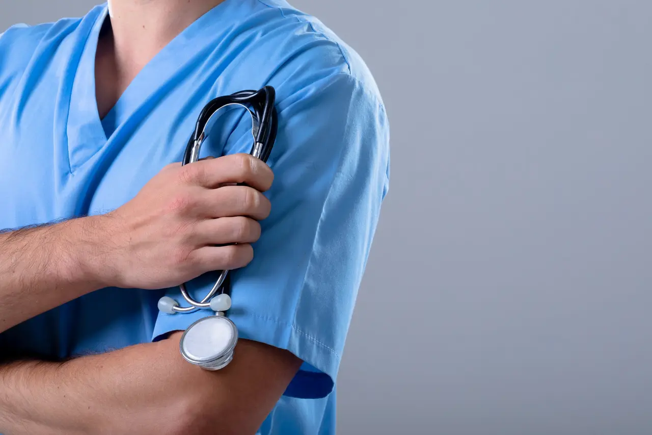 A practitioner in blue scrubs holding a stethoscope against a plain grey background