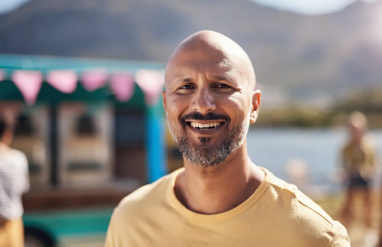 A smiling man in a yellow T-shirt outdoors by the water, with mountains behind him