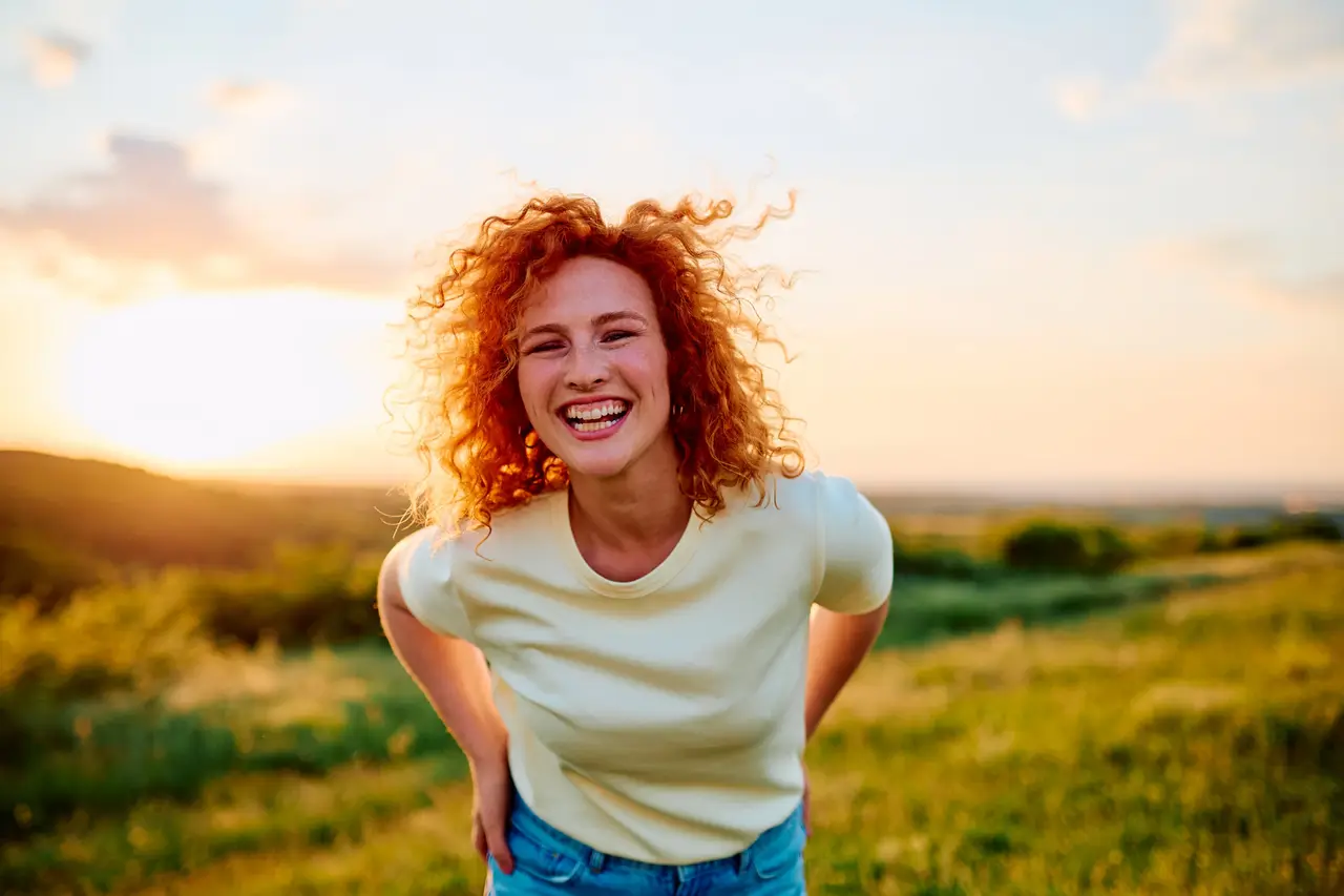 A laughing woman with red curls in a grassy field at sunset