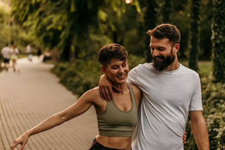 A couple laughing as they walk arm in arm through a leafy park